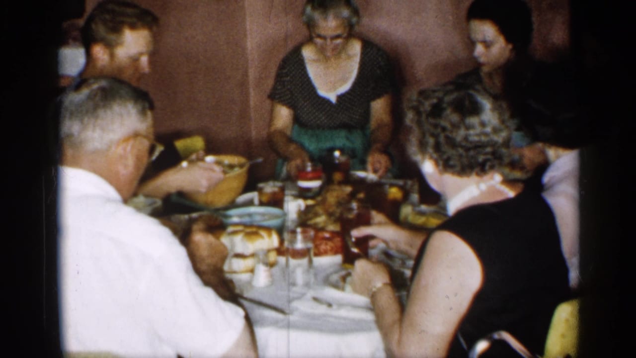 Adults seated around a dining table with plates, glasses, bread, and a large roasted bird.
