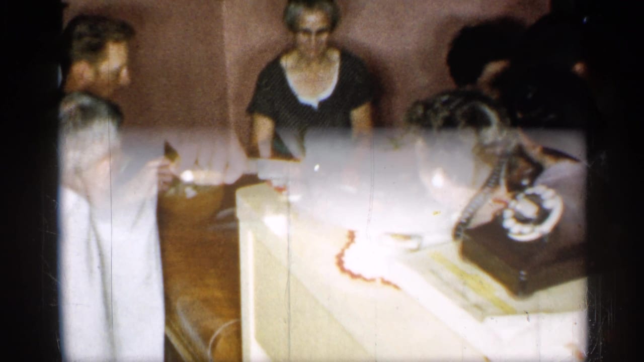 1955 New York home-movie frame with a frosted cake on a side table near a black rotary telephone.