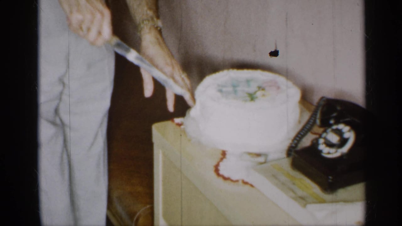 1955 New York home-movie frame showing a man cutting a white frosted cake beside a rotary telephone.