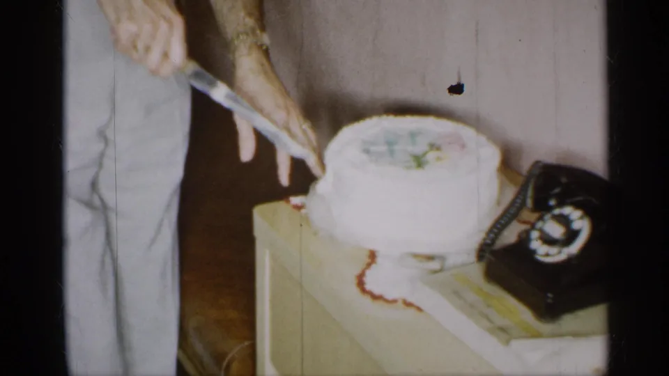 1955 New York home-movie frame showing a man cutting a white frosted cake beside a rotary telephone.