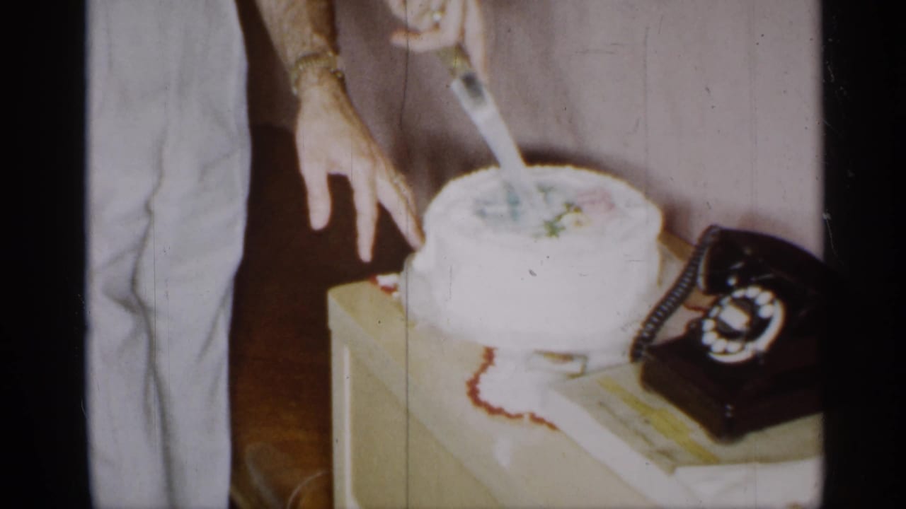 1955 New York home-movie frame of cake cutting in a private interior, with a rotary telephone on the same table.