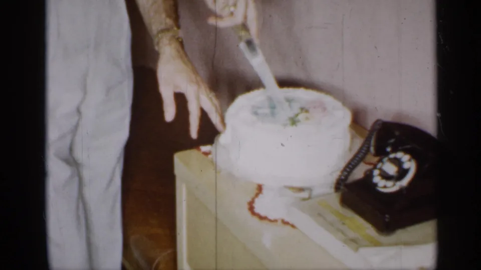1955 New York home-movie frame of cake cutting in a private interior, with a rotary telephone on the same table.