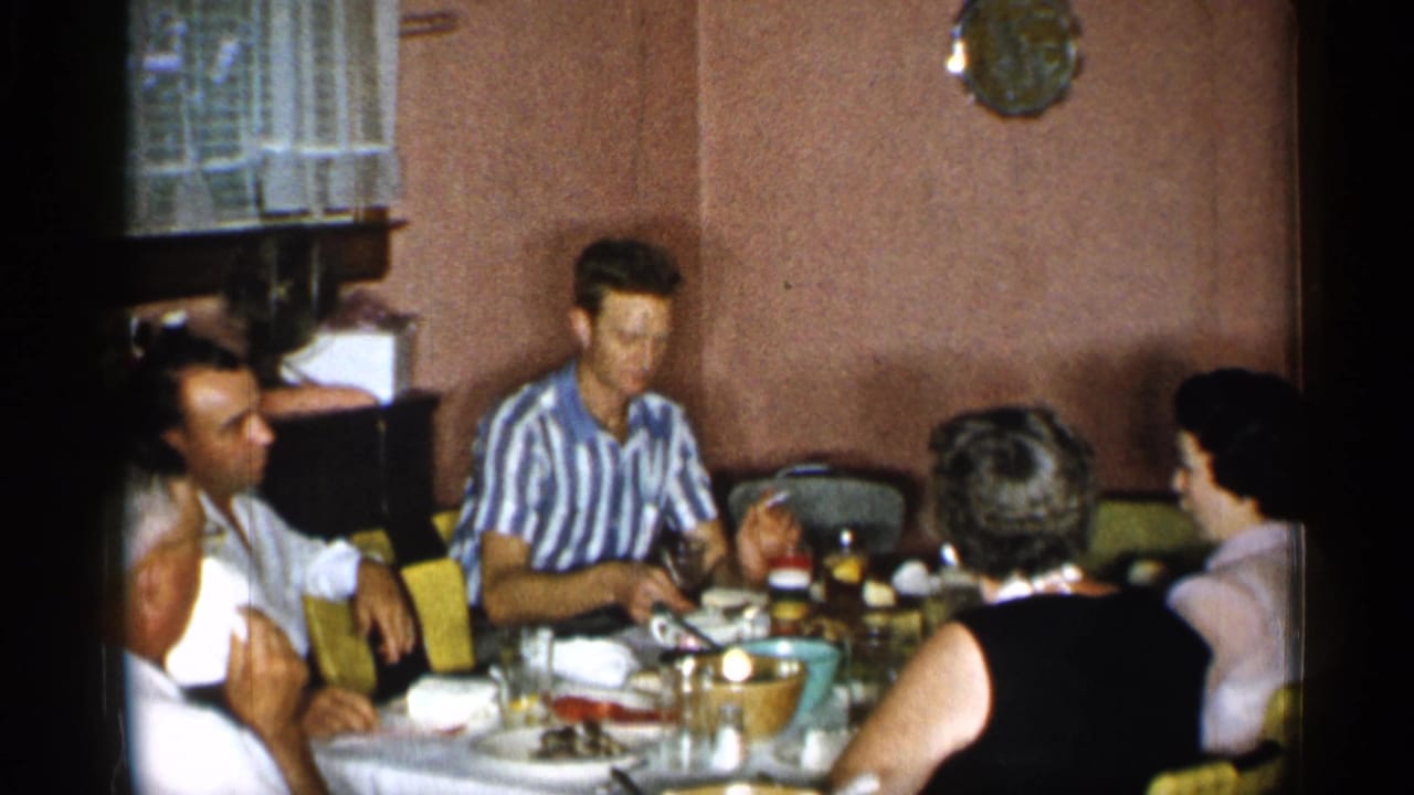 A man in a blue and white striped shirt sits at a 1955 New York dining table with adults, dishes, and glassware around him.