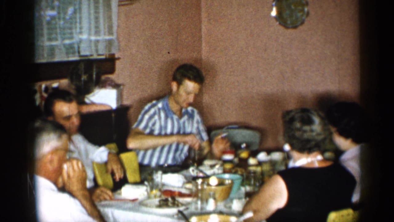 A man reaches toward the table during a 1955 New York family meal, with adults seated around dishes, glasses, and serving bowls.