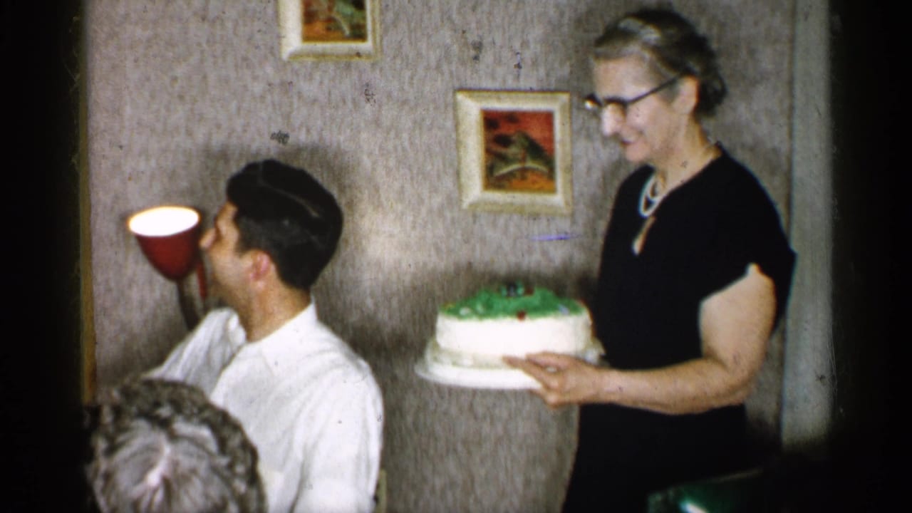 Woman in black dress holds a decorated cake beside a seated man at a dining table.