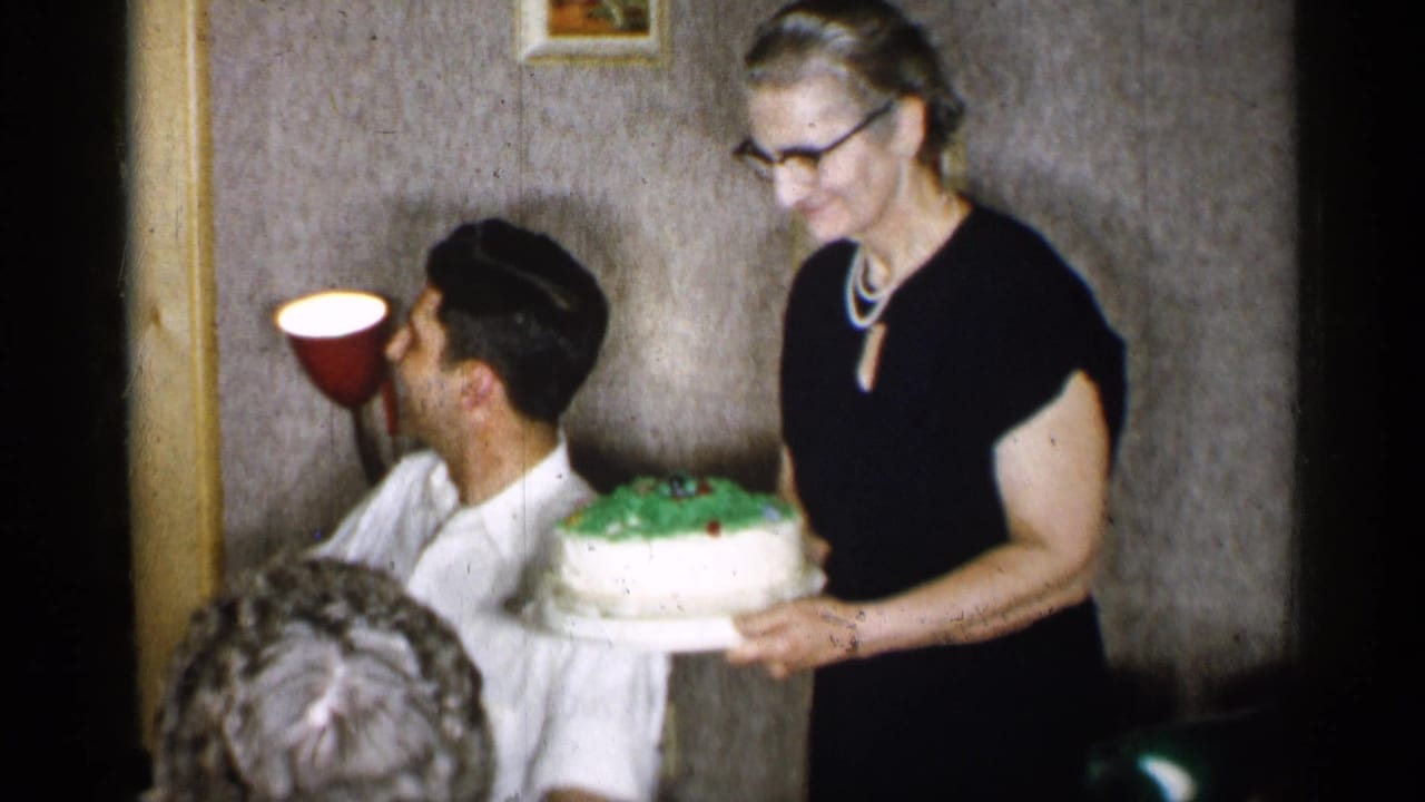 The woman in a black dress presents the decorated cake near the seated man, with wallpaper, small framed art, and a red wall lamp behind them.