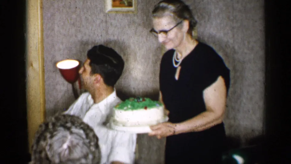 The woman in a black dress presents the decorated cake near the seated man, with wallpaper, small framed art, and a red wall lamp behind them.