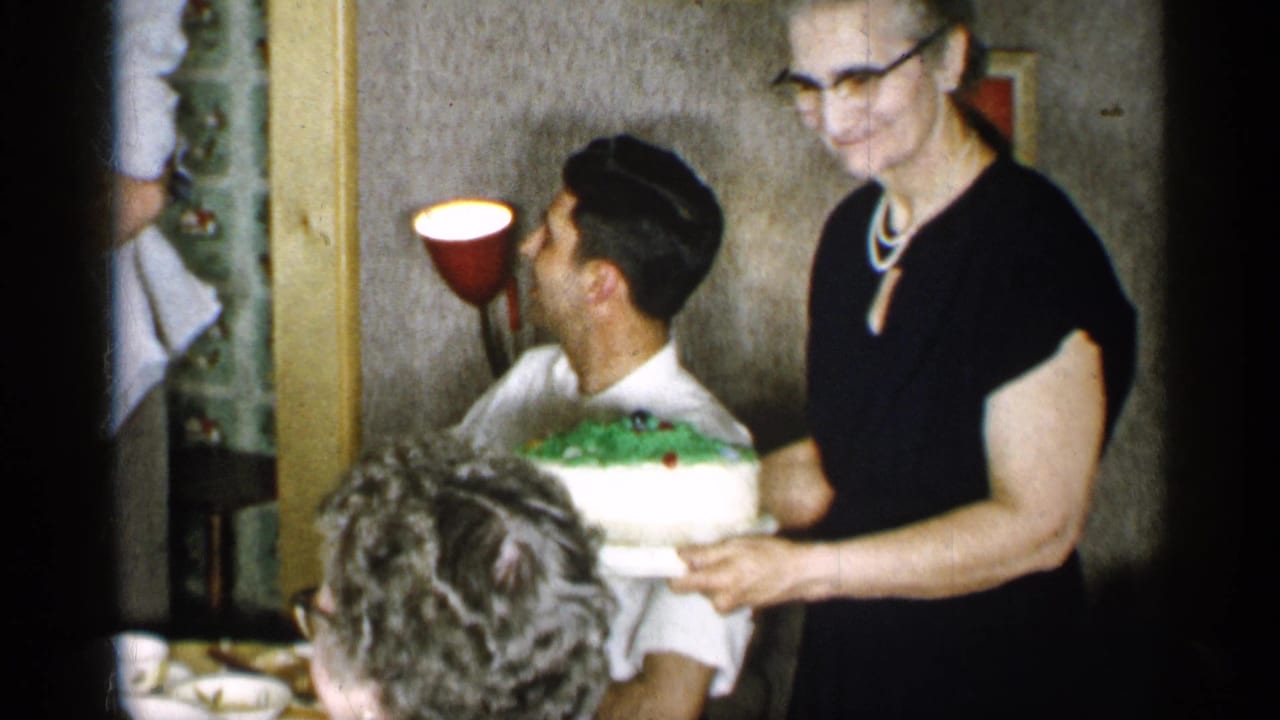 The woman moves the white and green cake closer to the table, with the seated man, dinnerware, and another person partly visible in the foreground.
