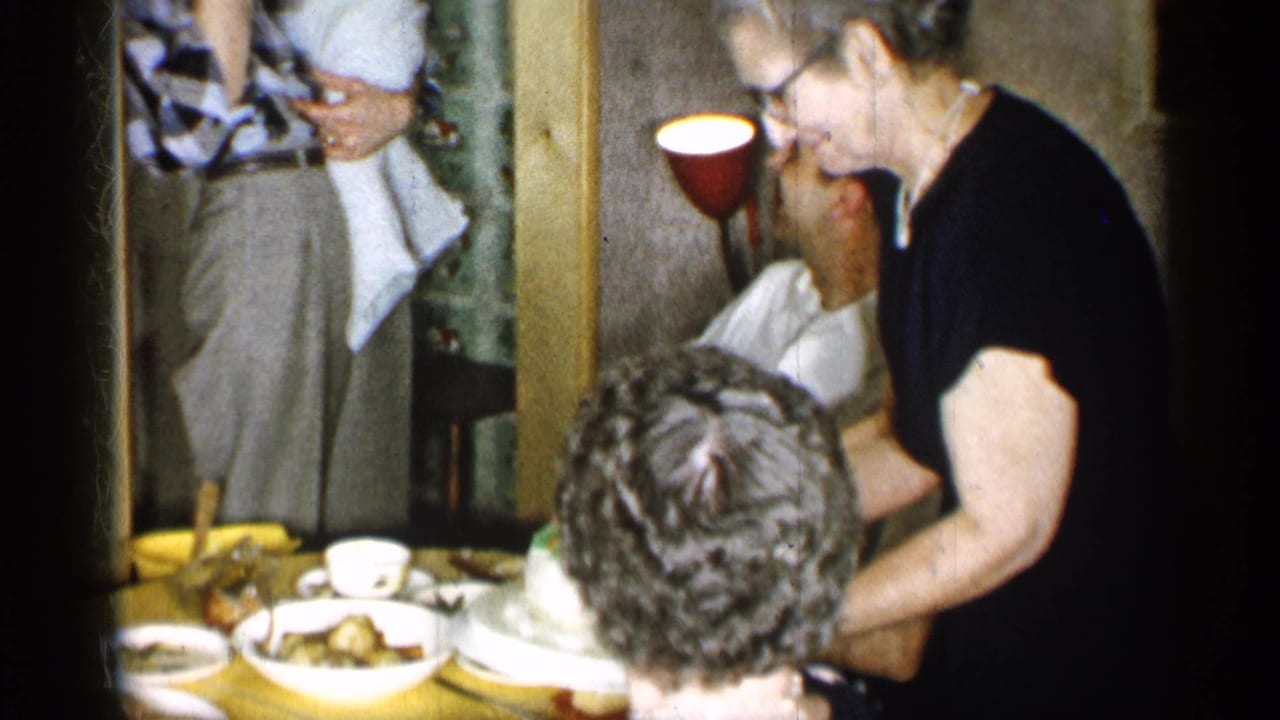 At the end of the clip, the woman leans over the dining table with the cake among plates, cups, and seated relatives in the private home.