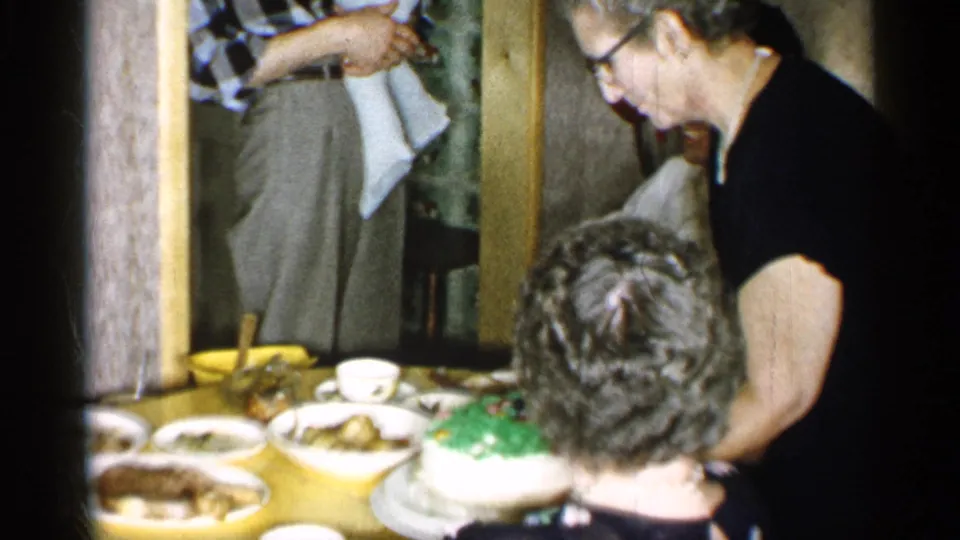 1955 New York home-movie frame showing a woman in a black dress serving near a green-and-white cake at an indoor dinner table.