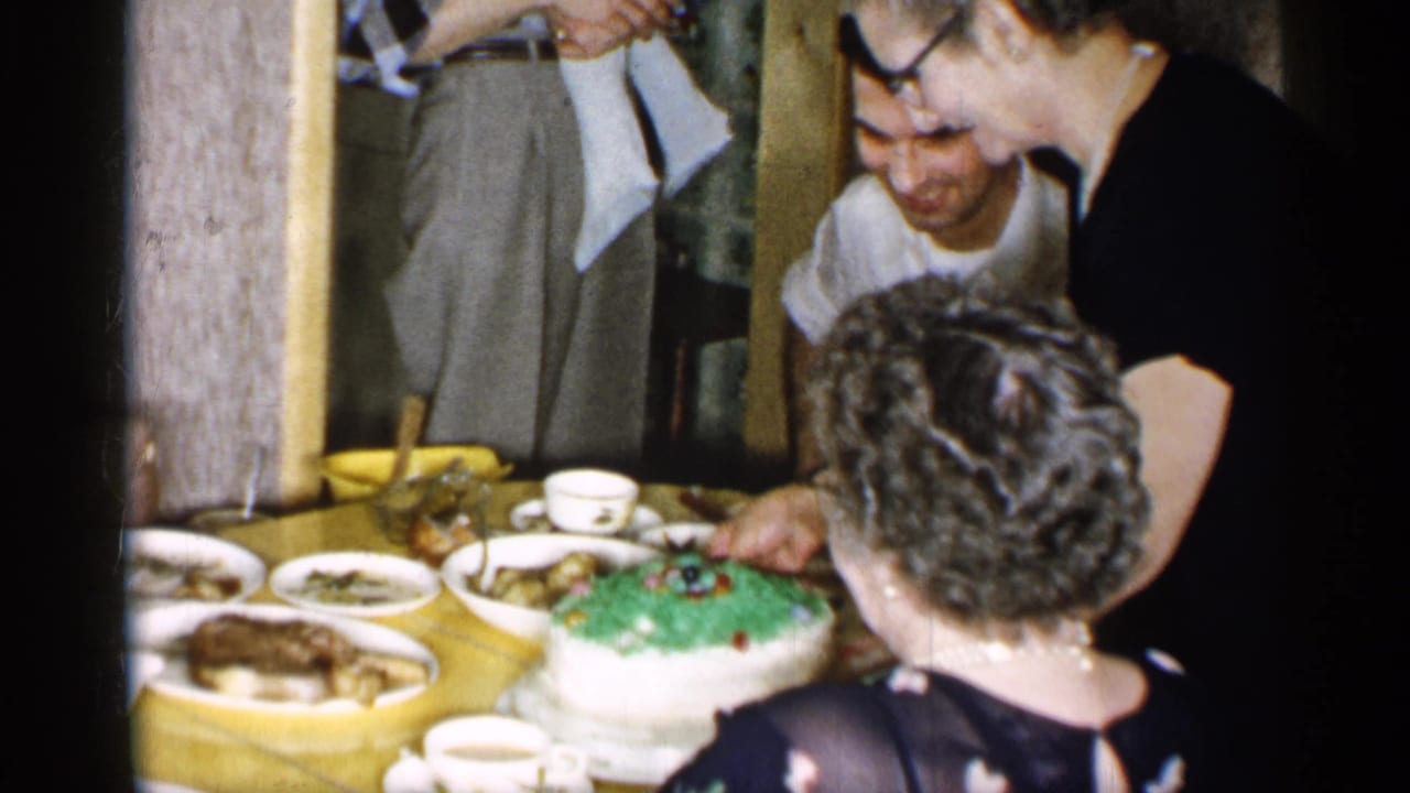 1955 New York home-movie frame with adults around a yellow-covered dinner table and a green-frosted cake in the center.