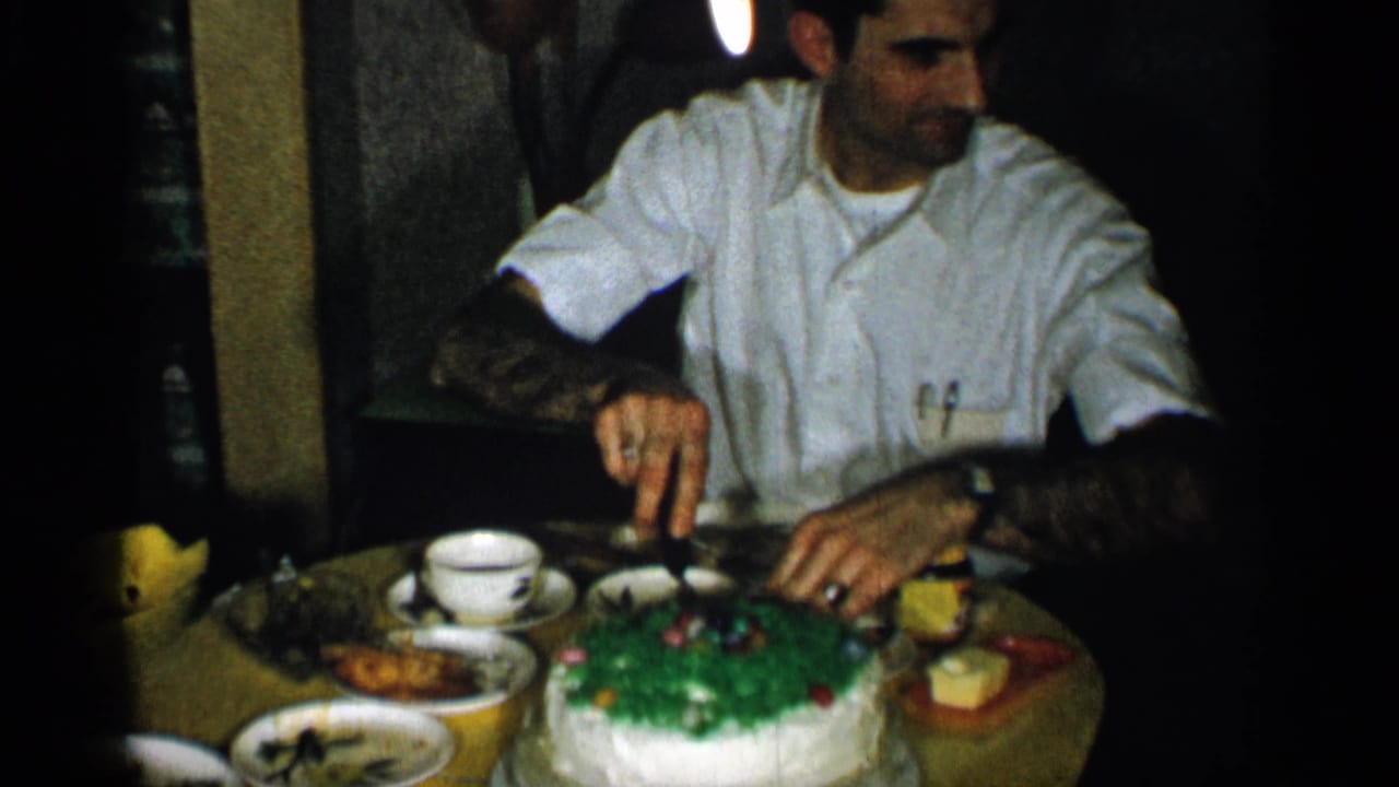 1955 New York home-movie frame showing a man in a white shirt cutting a green-and-white cake at a family dinner table.