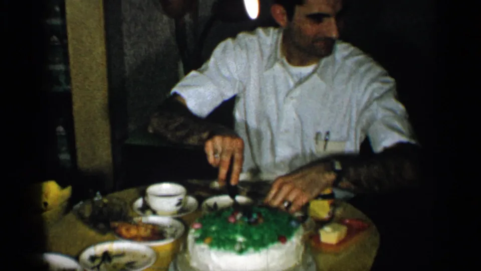 1955 New York home-movie frame showing a man in a white shirt cutting a green-and-white cake at a family dinner table.