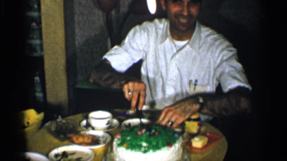 1955 New York home-movie frame showing a smiling man cutting a green-frosted cake during an indoor family meal.