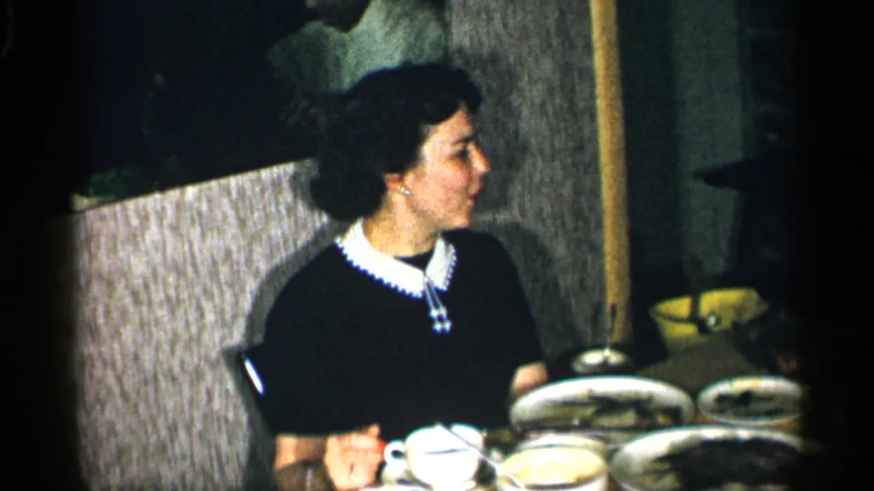 In supported 1955 New York home-movie footage, a woman in a black dress with a white collar sits at a restaurant table.