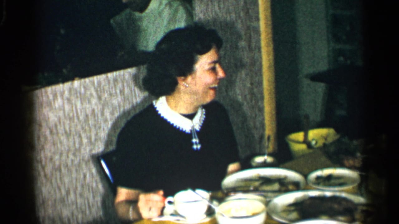 The woman remains seated at the table, looking toward someone off camera as plates and cups fill the foreground.