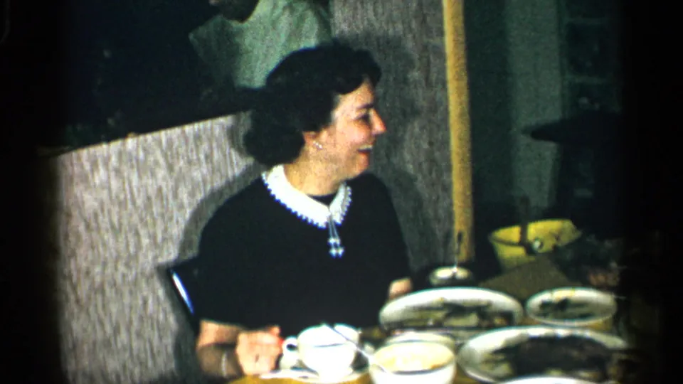 The woman remains seated at the table, looking toward someone off camera as plates and cups fill the foreground.