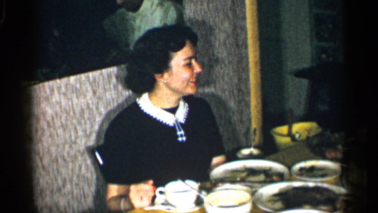 A woman smiles while seated at the restaurant table, with tableware and serving dishes visible in soft 8mm color.