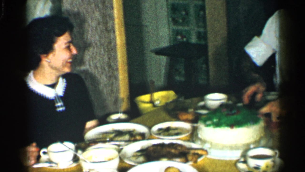 The table is more fully visible with plates, cups, serving dishes, a green-topped cake, and a companion partly entering the frame.