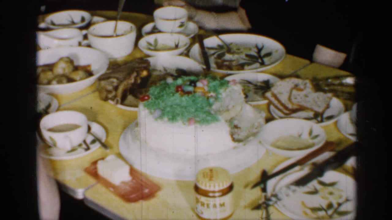 Close home-movie view of the 1955 New York dining table with a large decorated cake, sliced bread, cups, bowls, and serving dishes.