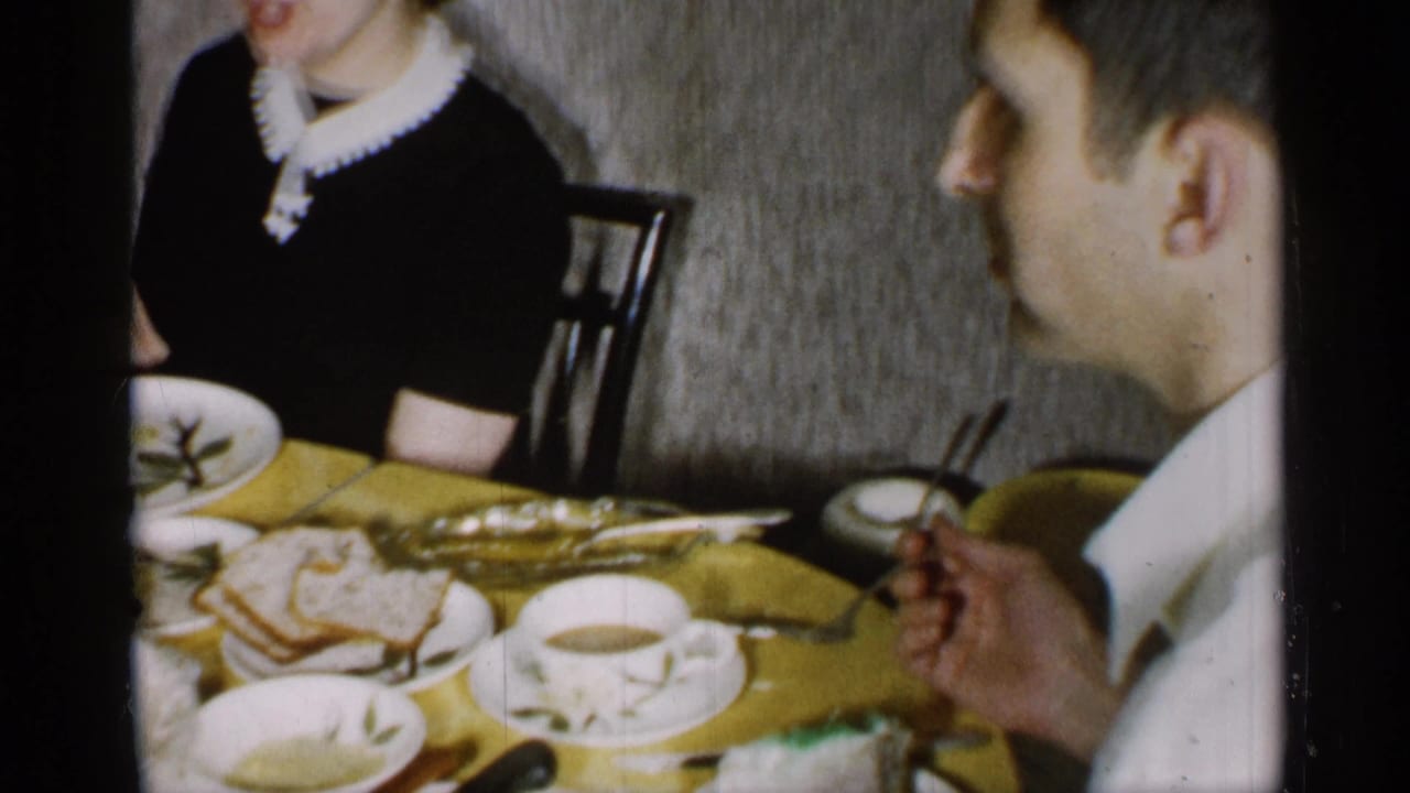 New York 1955 home-movie frame with a man seated in profile beside a dining table holding cups, bread, utensils, and a partially visible cake.