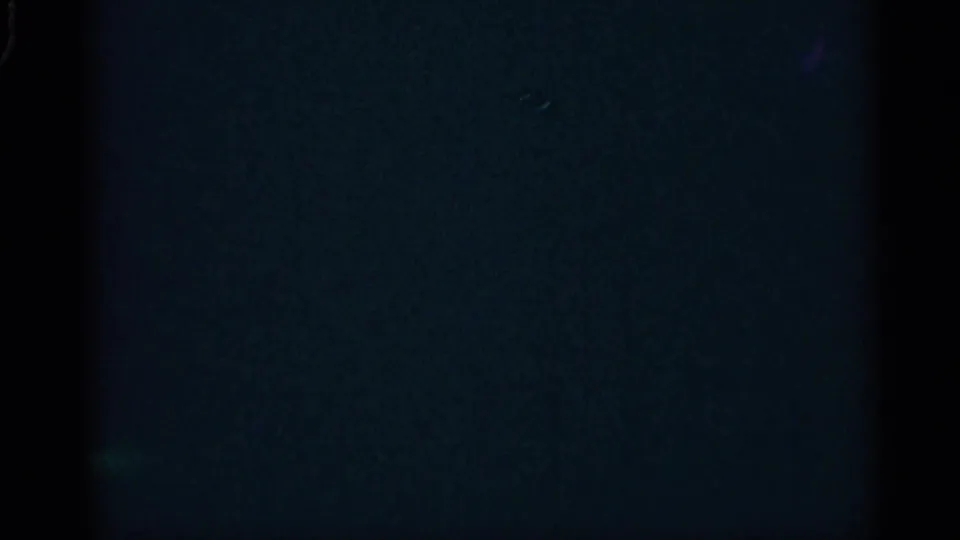 Dark 8mm leader-like frame with faint blue exposure, grain, and vertical edge shadows before the interior scene appears.