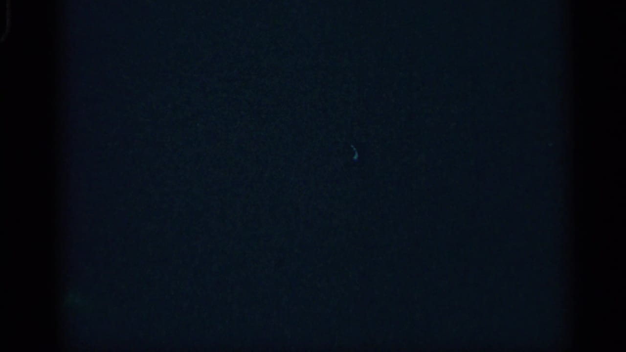 Mostly dark 8mm frame with faint bluish exposure, visible grain, and reel-edge shading, with no clear subject visible.