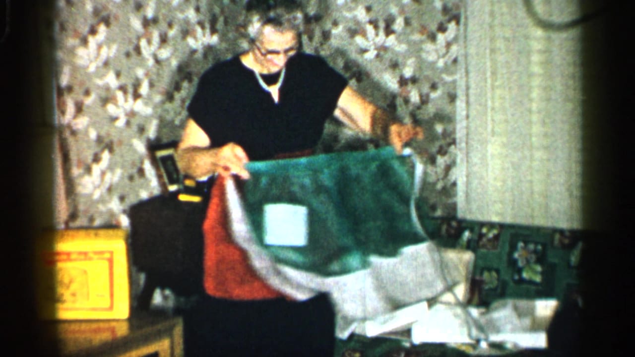 In a 1955 New York home interior, an elderly woman in a red apron holds up a green and white apron near floral wallpaper and papers.