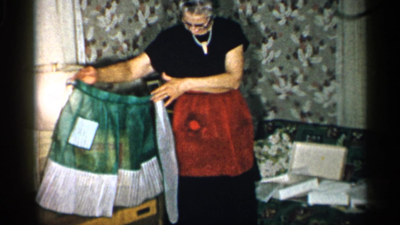 In a private New York interior from the supplied 1955 metadata, an elderly woman in a red apron examines a green apron by a sofa and scattered papers.