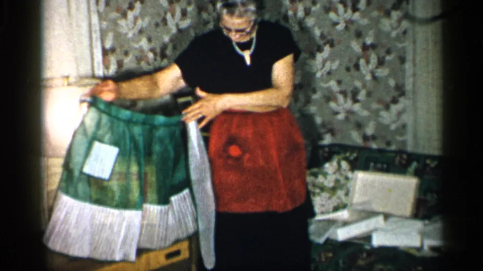 In a private New York interior from the supplied 1955 metadata, an elderly woman in a red apron examines a green apron by a sofa and scattered papers.