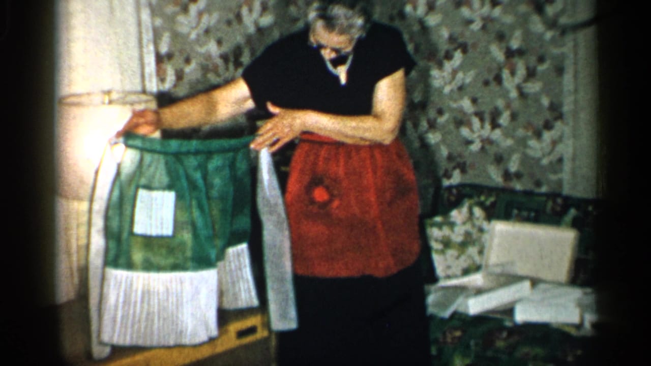 Woman in glasses and red apron holds up a green and white apron indoors.