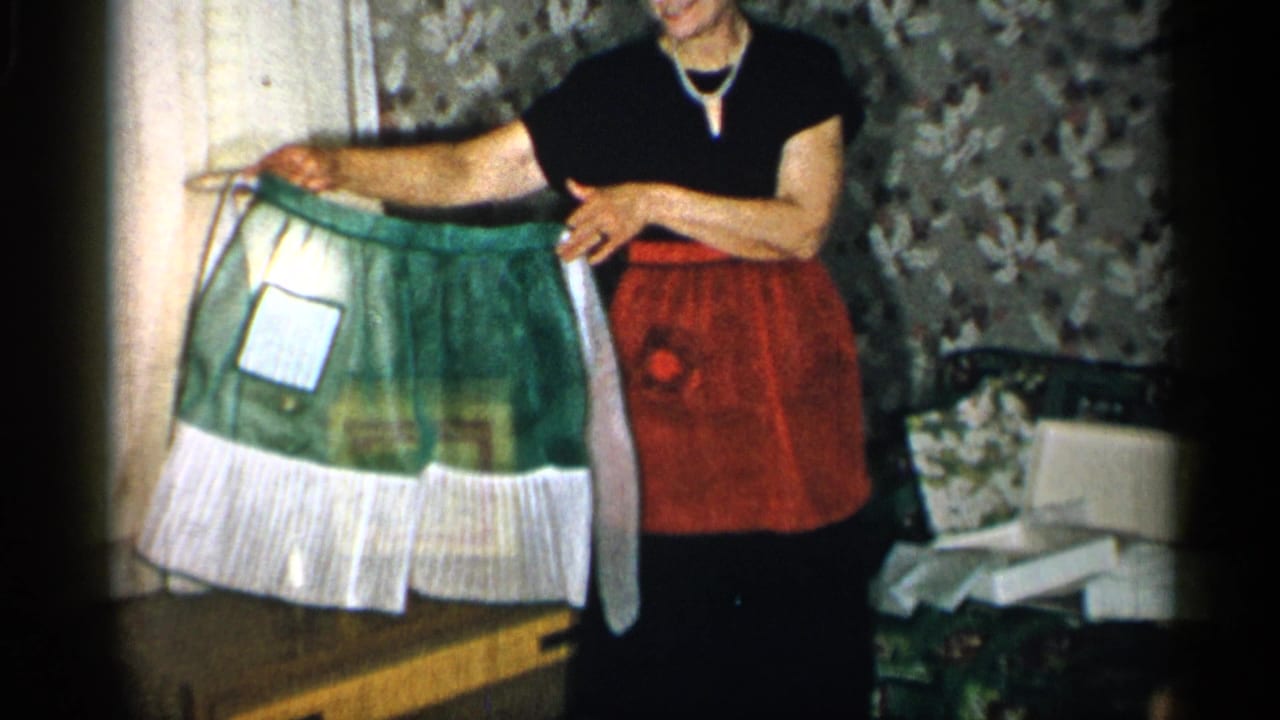 The woman stands in the private interior, holding the green-and-white apron open so its front panel and ruffled lower edge are visible.