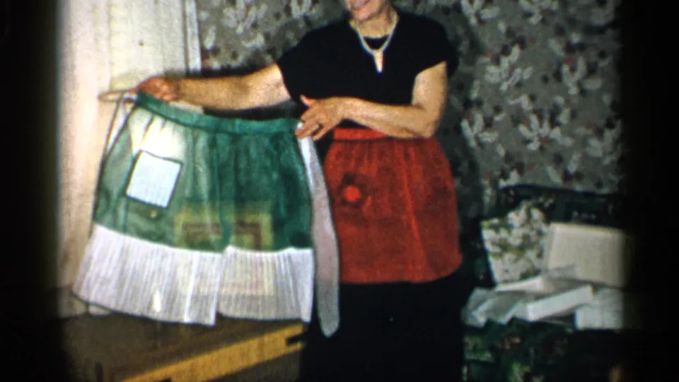 The woman stands in the private interior, holding the green-and-white apron open so its front panel and ruffled lower edge are visible.