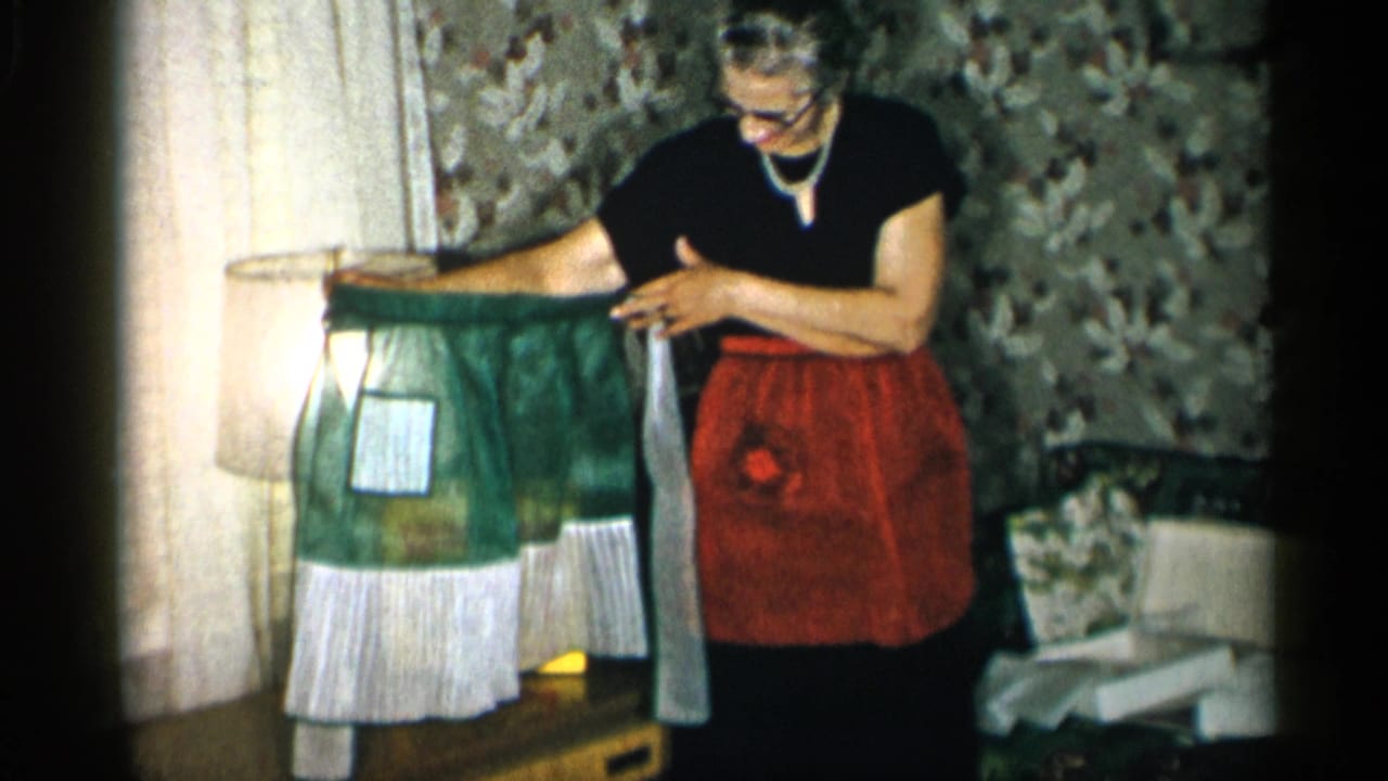 A woman in a black dress and red apron studies the green apron, with a white rectangular patch and ruffled hem visible near the lamp.