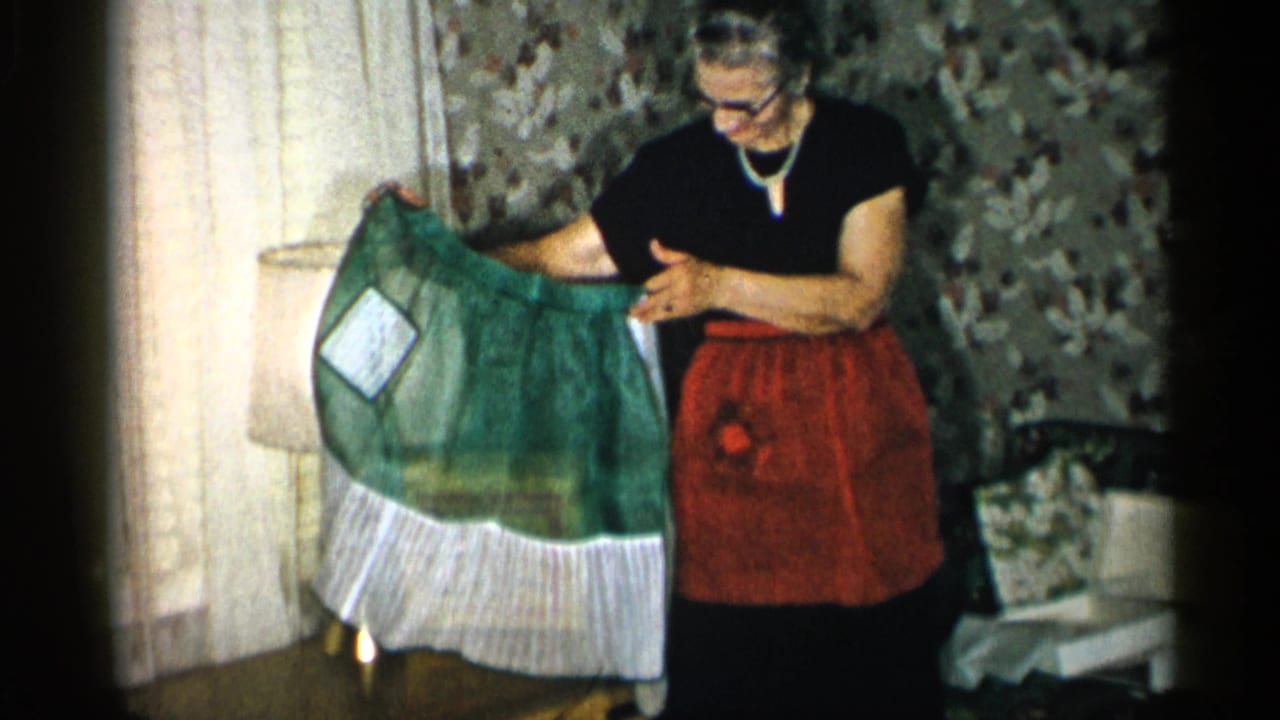 The woman angles the green apron toward the camera, showing its sheer fabric, white patch, and ruffled hem inside the New York home interior.