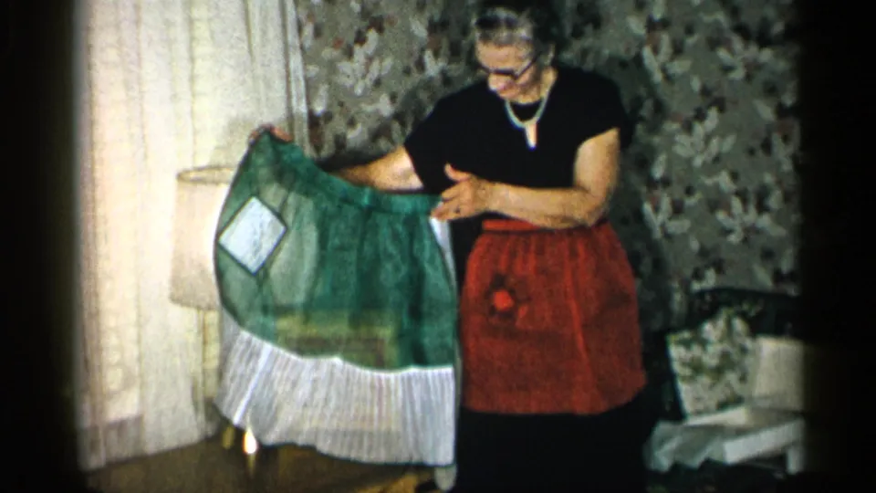 The woman angles the green apron toward the camera, showing its sheer fabric, white patch, and ruffled hem inside the New York home interior.