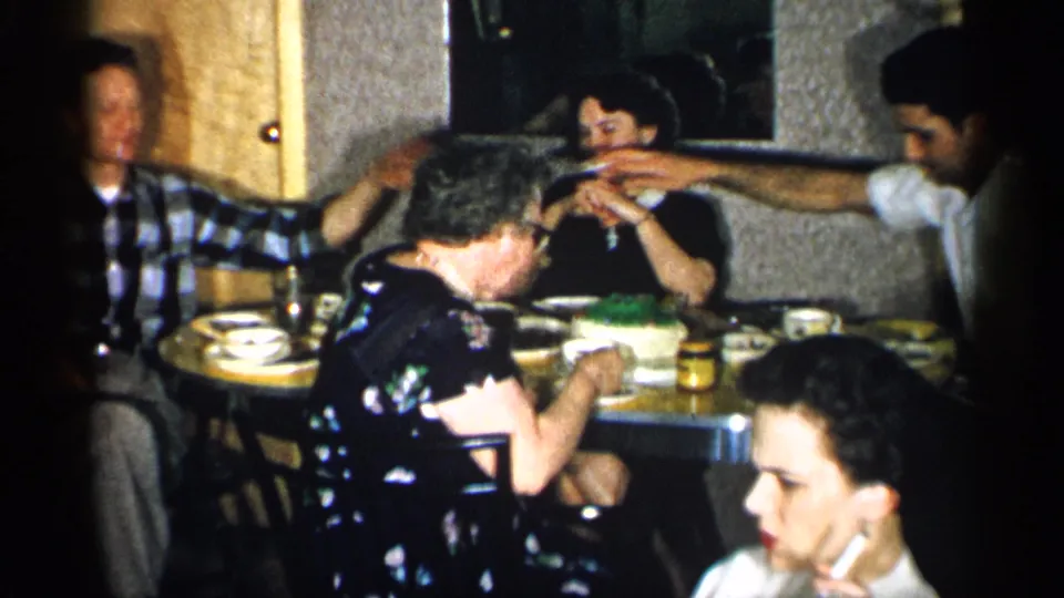 1955 New York home-movie frame showing adults seated around a dinner table, with a cake, cups, plates, and conversation visible.