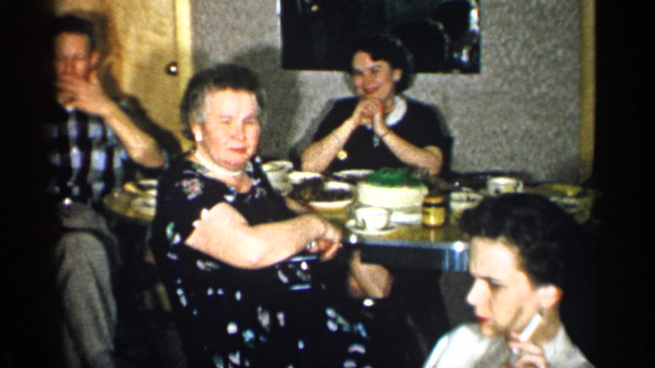 1955 New York home-movie frame with a woman in a dark floral dress near the table while other adults sit, eat, and talk.