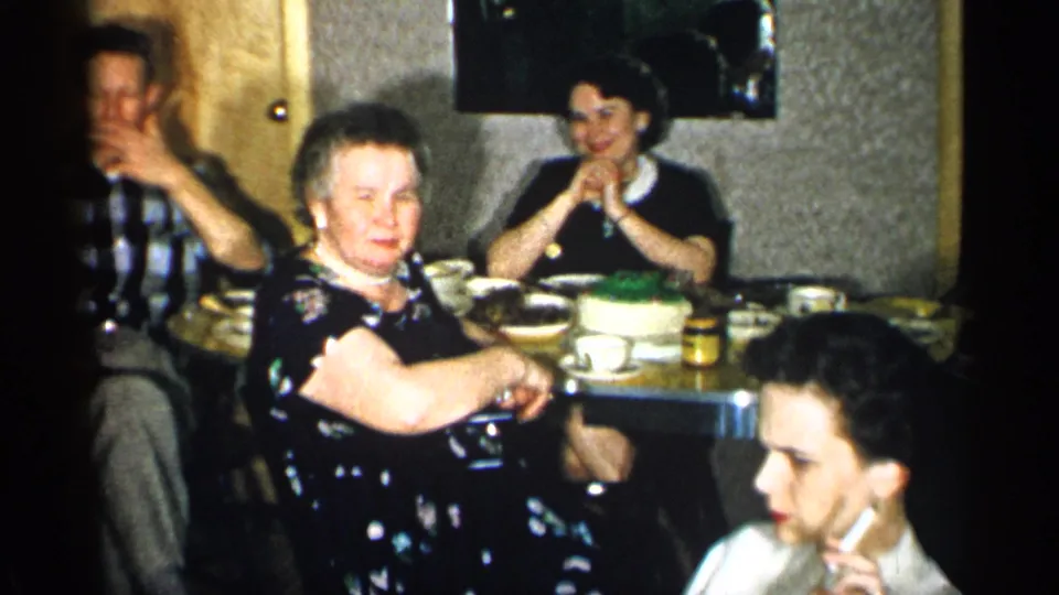1955 New York home-movie frame with a woman in a dark floral dress near the table while other adults sit, eat, and talk.