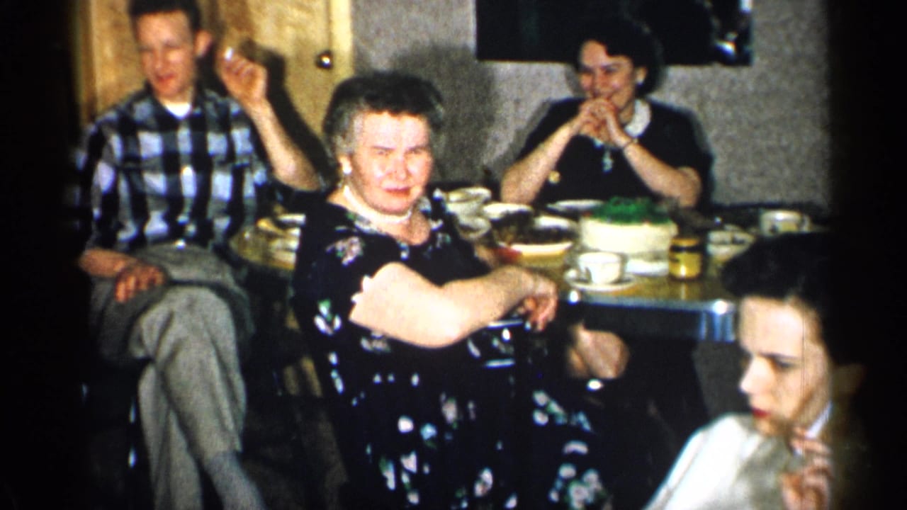 1955 New York home-movie frame showing adults at a family table, including a man in plaid and a woman in a dark floral dress.