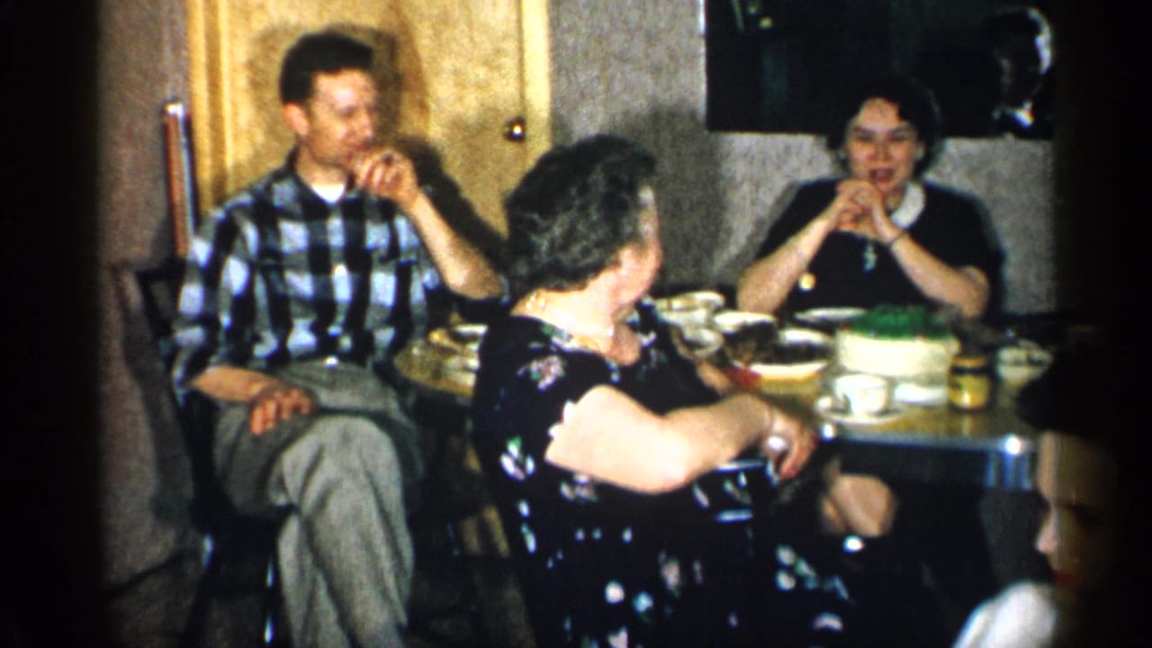 1955 New York home-movie frame of adults talking around a small dinner table in a private interior with tableware and dessert.