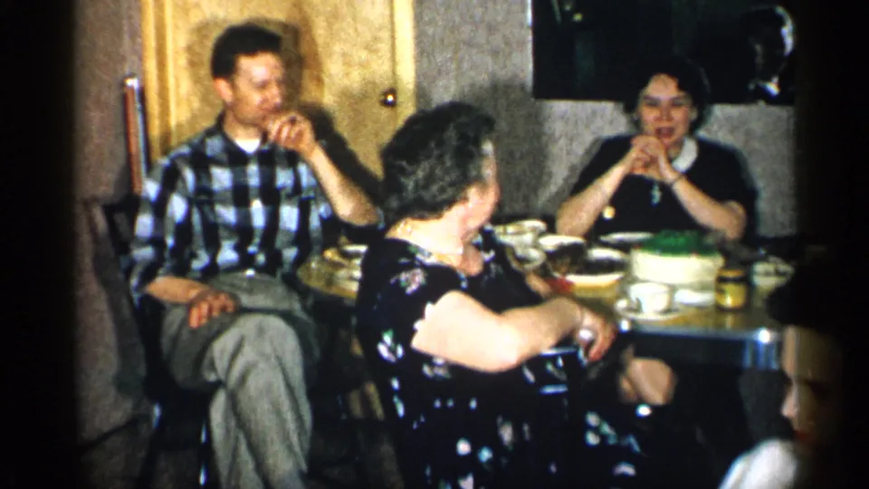 1955 New York home-movie frame of adults talking around a small dinner table in a private interior with tableware and dessert.