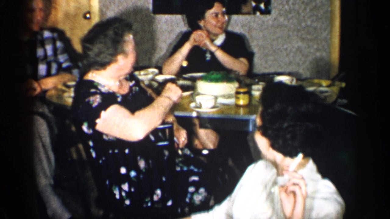 1955 New York home-movie frame of adults gathered in a restaurant booth, with a woman in a white top smoking in the foreground.
