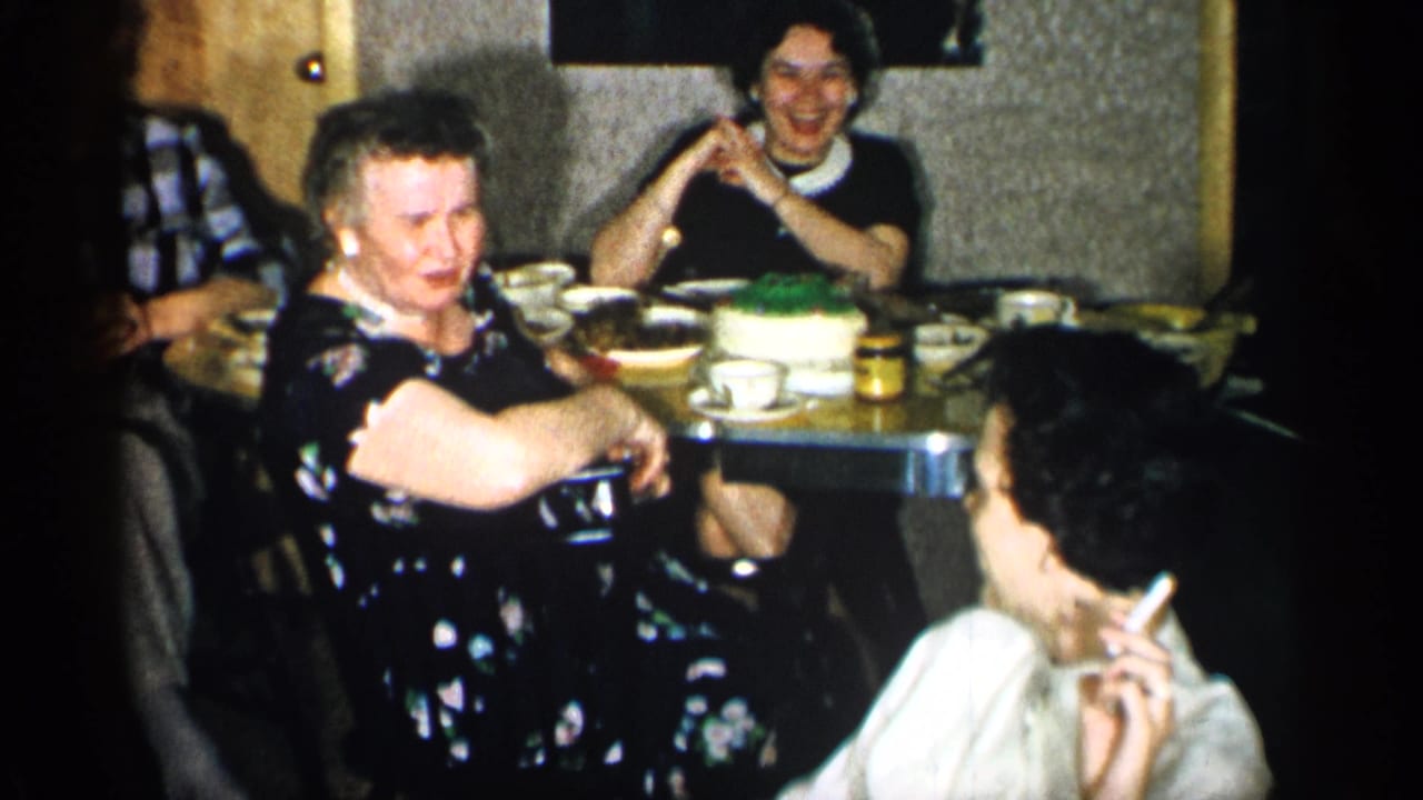 1955 New York home-movie frame showing a laughing woman in a dark floral dress at a table with cups, jars, and dessert nearby.