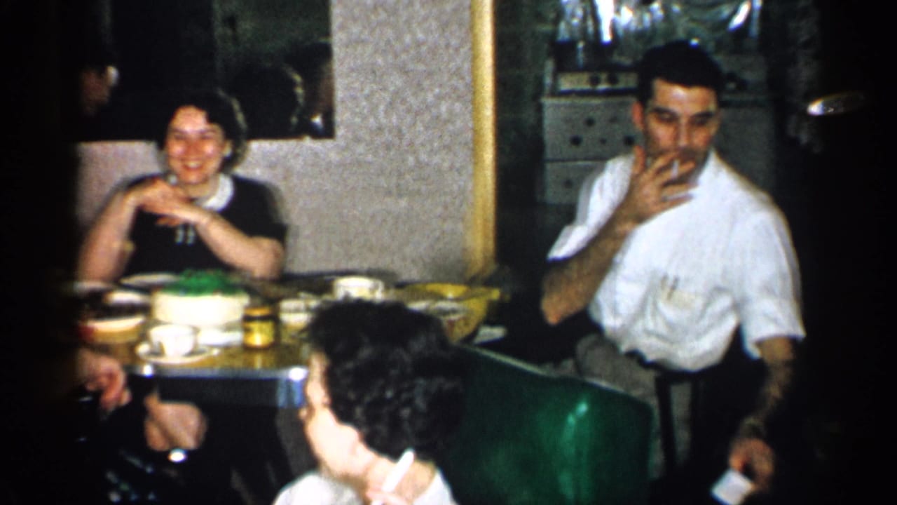 1955 New York home-movie frame with adults in a restaurant interior, including a seated man in a white shirt smoking near the table.