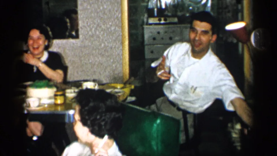 1955 New York 8mm frame of adults at a dim restaurant table with a cake as a man in a white shirt gestures toward the camera.