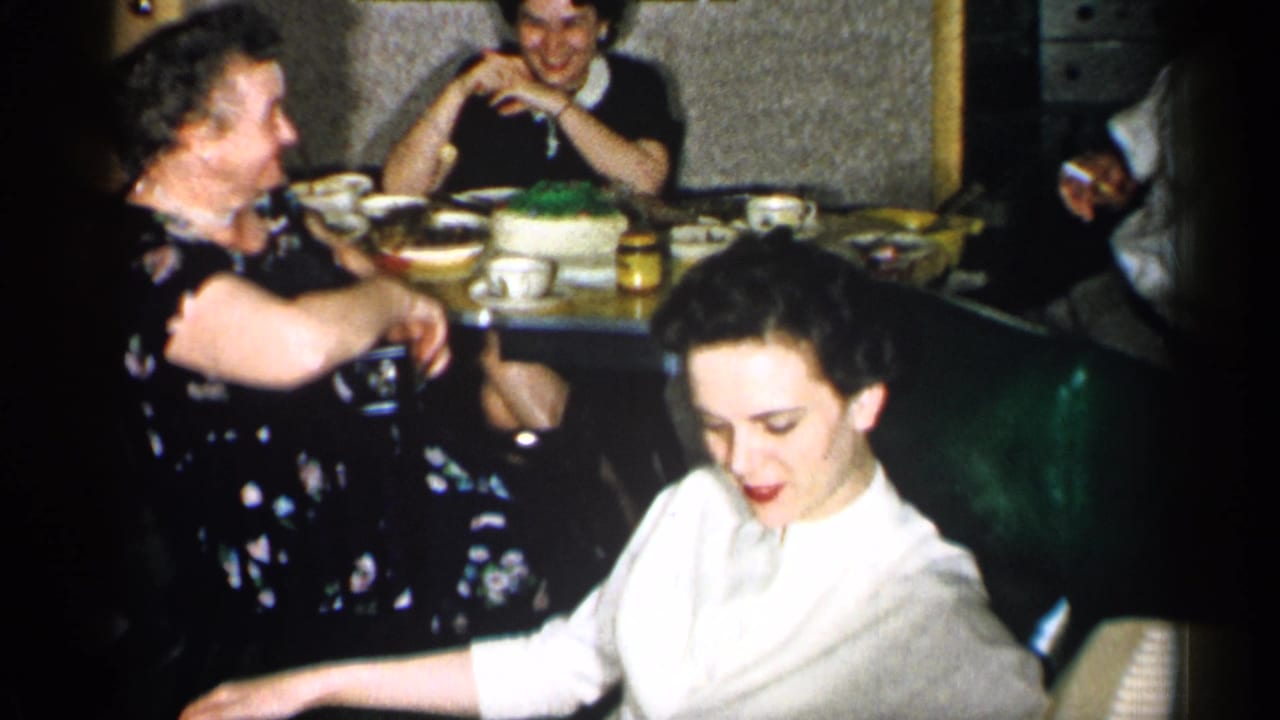 1955 New York home-movie frame of women at a restaurant booth, with cake, cups, and a foreground figure in soft focus.