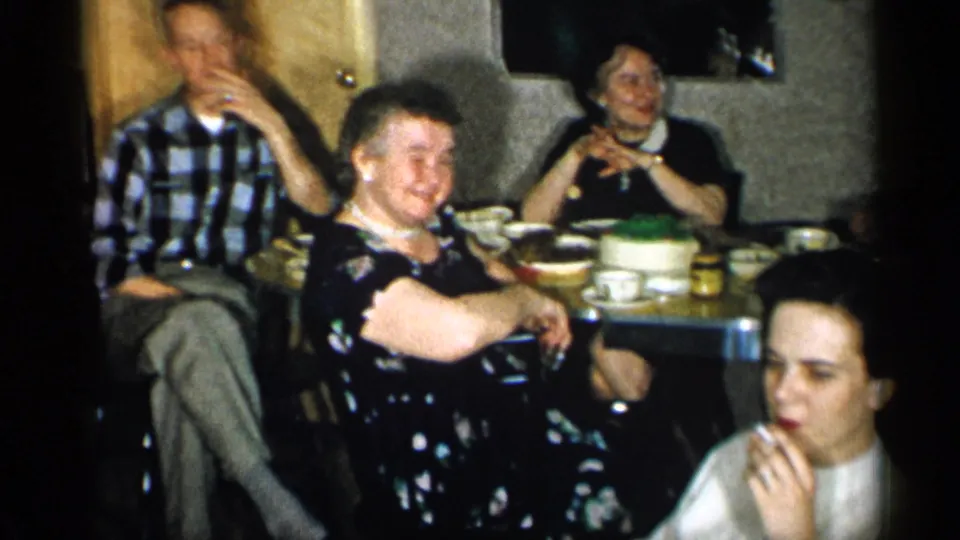 1955 New York restaurant gathering with adults seated around a table, a cake at center, and a woman in front eating or touching her mouth.