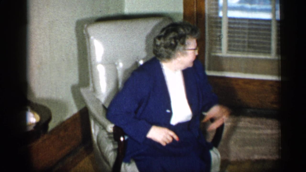 Older woman in a navy suit seated in a light armchair beside wood-framed windows.