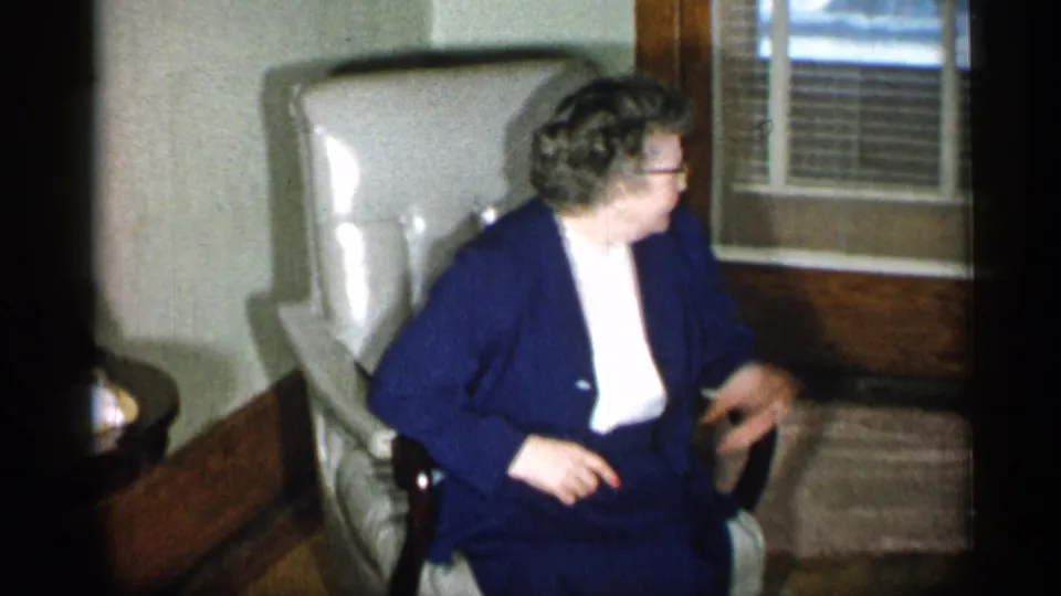In a 1955 New York home-movie frame, an older woman in glasses and a navy suit sits in a light armchair near venetian blinds.